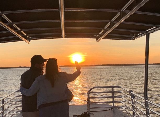 Silhouetted couple embracing on a covered boat deck taking a sunset selfie as the golden sun reflects across calm water