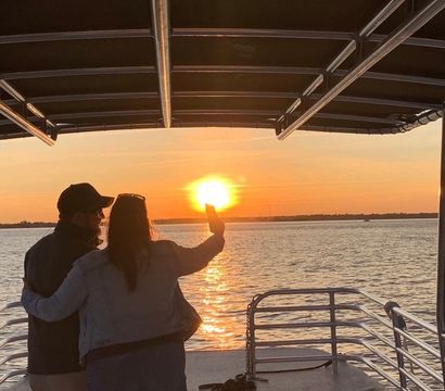Silhouetted couple embracing on a covered boat deck taking a sunset selfie as the golden sun reflects across calm water