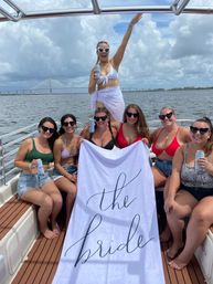 Boat bachelorette celebration: bride in white sarong and heart sunglasses raises her arm while friends in swimsuits hold a towel that reads 'the bride', canned drinks, waterfront with cable-stayed bridge and cloudy sky.