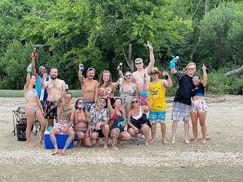 Large group of people on a sandy, tree-lined shore raising drinks and smiling at a casual summer beach party in swimsuits.