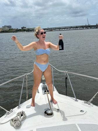 Smiling woman in a blue gingham bikini cheering with a bottle of champagne on the bow of a sailboat in a marina with a bridge and waterfront buildings in the background.