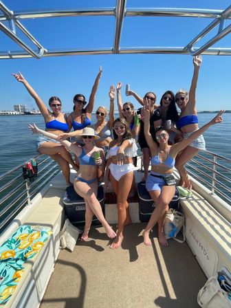 Group of women in swimsuits and sunglasses partying on a pontoon boat, arms raised and holding drinks under a bright blue summer sky with calm harbor water and a distant shoreline.