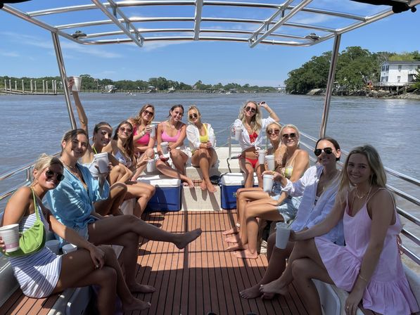 Group of women in swimsuits holding drinks on a sunny pontoon boat cruising a scenic river past docks and shoreline homes.