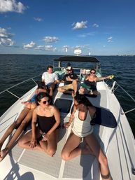 Friends relaxing with drinks on the bow of a yacht on a sunny day, lounging on deck cushions with blue sky, scattered clouds and calm open water in the background.