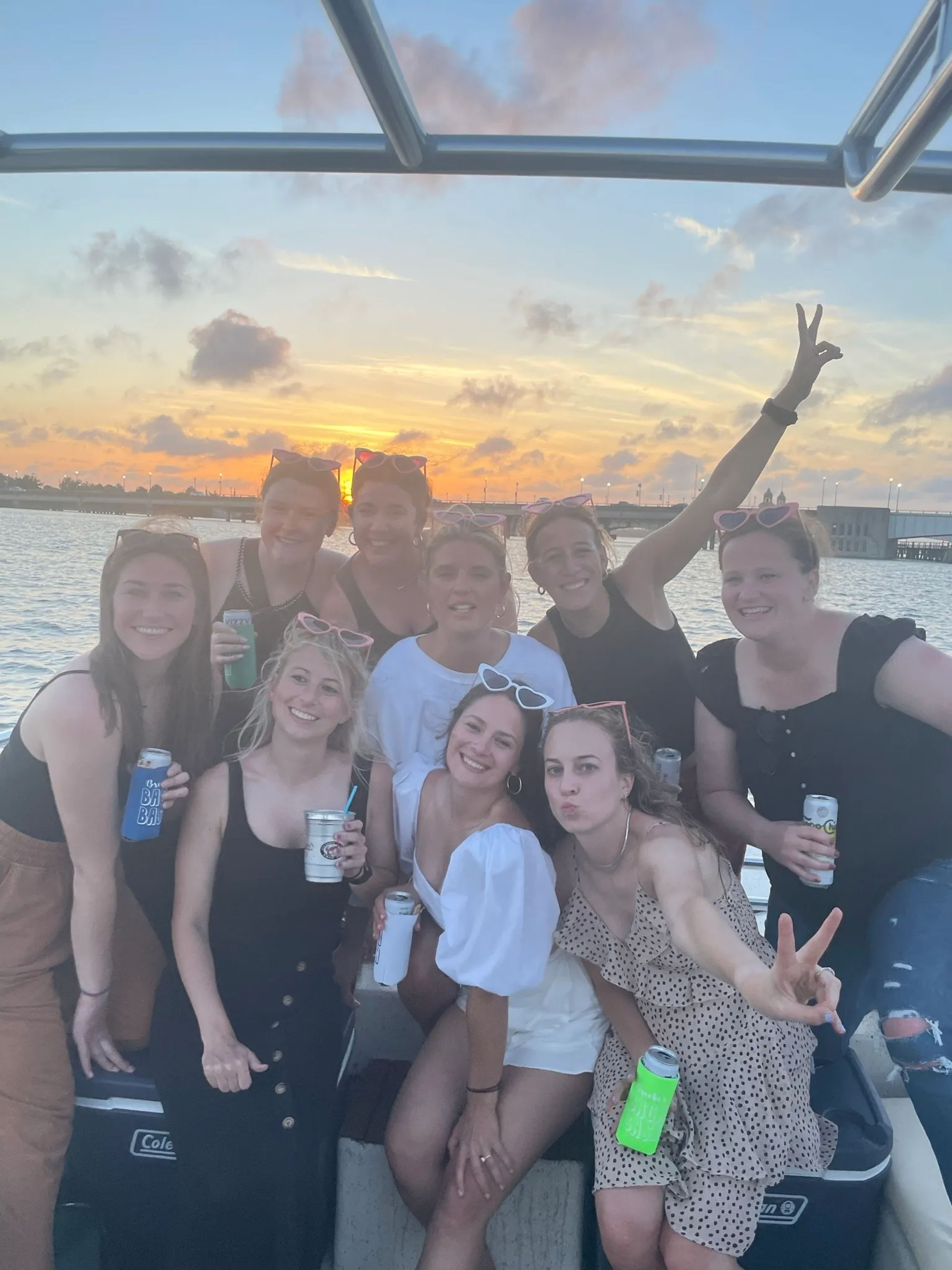Group of friends on a boat at sunset over a coastal harbor with a bridge in the background, holding drinks and posing for a fun waterfront photo
