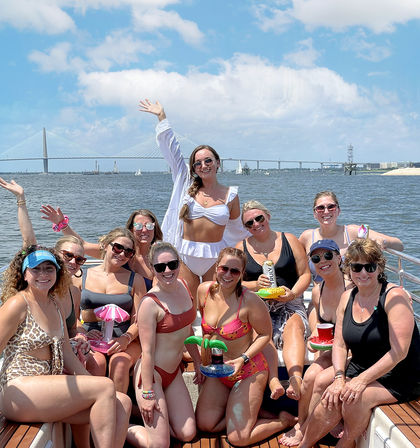 Cheerful group of women in colorful swimsuits posing and waving on a sunlit boat party, holding inflatable drink holders with sailboats and a cable-stayed bridge on the coastal harbor under a blue sky.