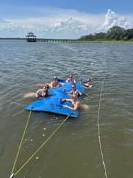 Six friends lounging on a blue tethered floating mat in a calm coastal bay, holding drinks near a wooden pier and gazebo with marsh grasses and puffy clouds on a sunny summer day