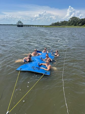 Six friends lounging on a blue tethered floating mat in a calm coastal bay, holding drinks near a wooden pier and gazebo with marsh grasses and puffy clouds on a sunny summer day