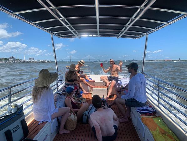 Group of people relaxing on a canopy-covered pontoon boat, cruising past a coastal city and suspension bridge on a sunny day, one person waving with a red cup.