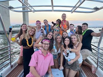Group of people on a lively party boat at sunset, wearing leis and holding drinks under a metal canopy with a bridge and harbor in the background.