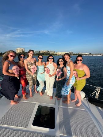 Nine friends barefoot on a yacht deck posing for a sunny summer boat-party group photo by a coastal skyline, wearing dresses and sunglasses and holding oversized face cutouts on sticks.