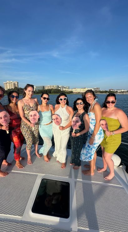 Nine friends barefoot on a yacht deck posing for a sunny summer boat-party group photo by a coastal skyline, wearing dresses and sunglasses and holding oversized face cutouts on sticks.