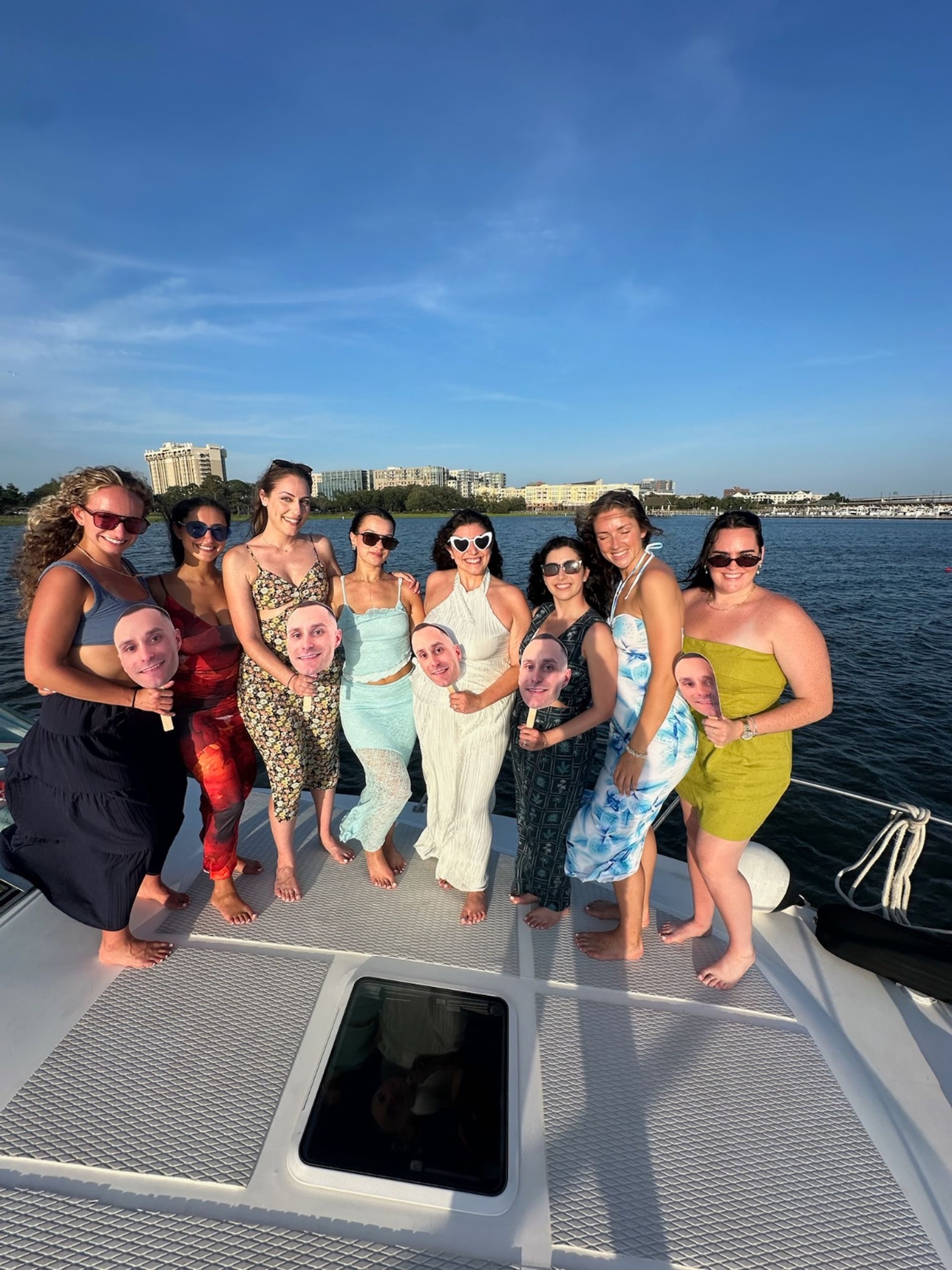 Nine friends barefoot on a yacht deck posing for a sunny summer boat-party group photo by a coastal skyline, wearing dresses and sunglasses and holding oversized face cutouts on sticks.