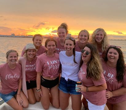 Group of ten smiling women on a boat at sunset, many in matching pink t‑shirts and casual shorts with one in a white tee, golden sky and waterfront skyline in the background.