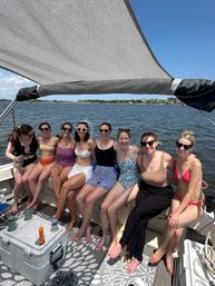 Eight friends in colorful swimsuits sitting on a shaded pontoon boat, laughing together with a coastal bay, waterfront homes and clear blue sky in the background on a sunny summer day.