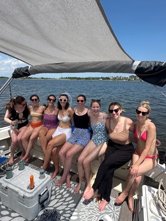 Eight friends in colorful swimsuits sitting on a shaded pontoon boat, laughing together with a coastal bay, waterfront homes and clear blue sky in the background on a sunny summer day.