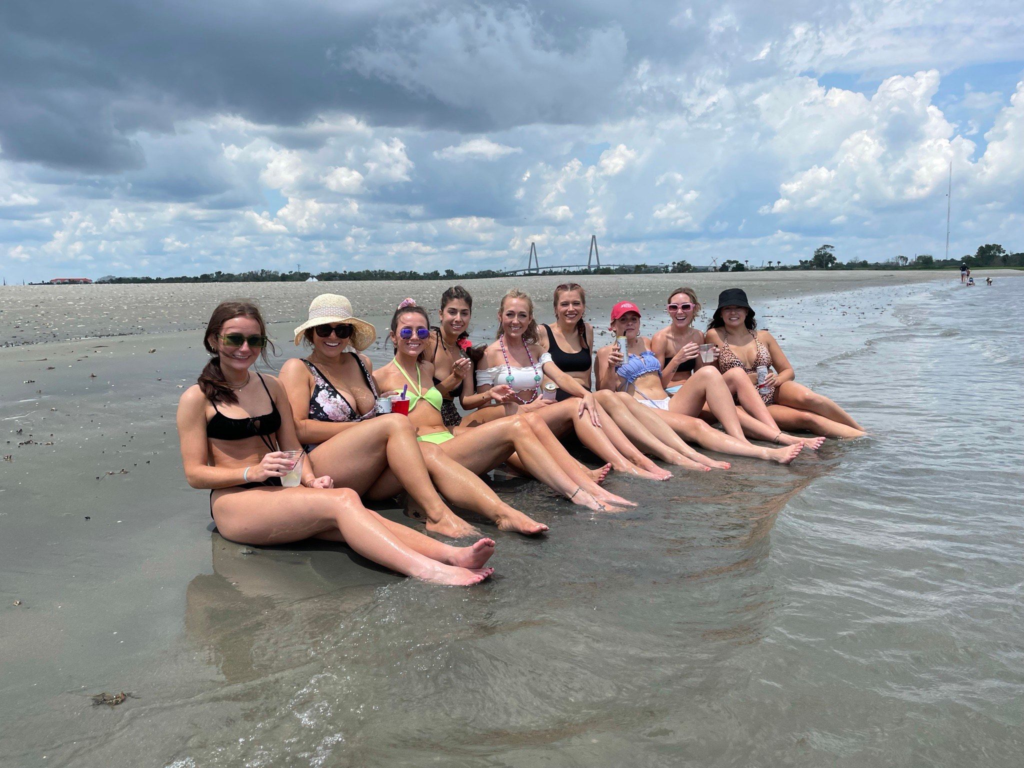 Group of friends in bikinis sitting with legs outstretched in shallow water on a sandy beach, holding drinks under a dramatic cloudy sky with a distant bridge on the horizon — fun summer coastal scene.