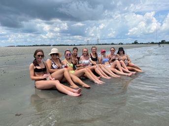 Group of friends in bikinis sitting with legs outstretched in shallow water on a sandy beach, holding drinks under a dramatic cloudy sky with a distant bridge on the horizon — fun summer coastal scene.
