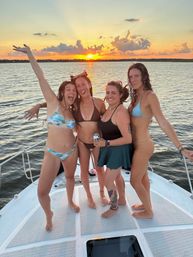 Four friends in swimsuits smiling and posing on a boat deck during a golden sunset over calm coastal waters.