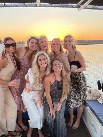 Seven women smiling with drinks on a boat at golden sunset over calm water — relaxed sunset cruise vibe.