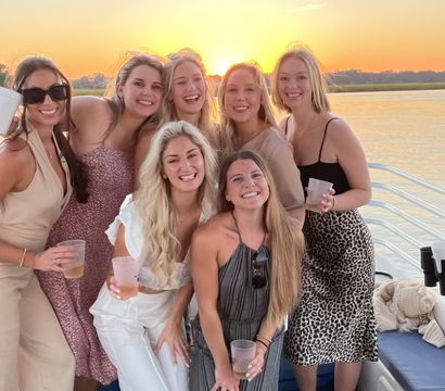 Group of friends smiling on a boat deck with drinks as a golden sunset glows over calm water — sunset cruise vibes