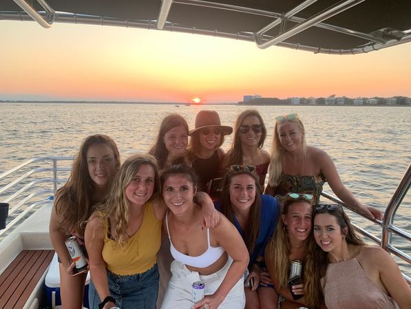Nine friends on a sunset boat cruise, smiling on deck with drinks against calm bay waters and a coastal skyline at golden hour.