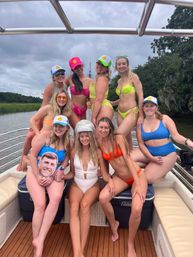 Group of women in vibrant swimsuits and smiley-logo trucker hats posing on a boat deck by a marshy river under a cloudy sky; one wears a rhinestone tiara and another holds a face cutout for a playful summer boat outing.