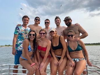 Eight friends in swimsuits smiling and posing on a boat near a sandy shoreline at a coastal bay, seagulls overhead and a cloudy summer sky