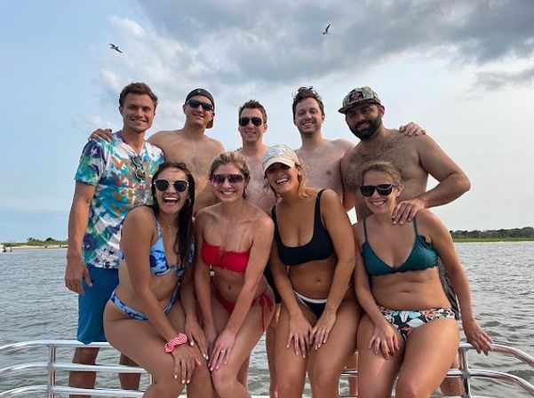 Eight friends in swimsuits smiling and posing on a boat near a sandy shoreline at a coastal bay, seagulls overhead and a cloudy summer sky