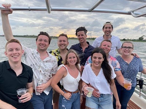 Group of friends smiling and holding drinks on a sunset boat cruise over calm water, cloudy sky and distant shoreline in the background.