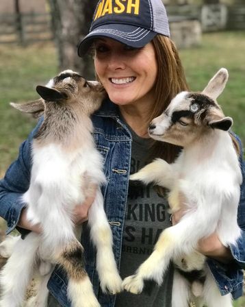 Smiling woman in a denim jacket and baseball cap holding two baby goats on a rural farm, one nuzzling her cheek