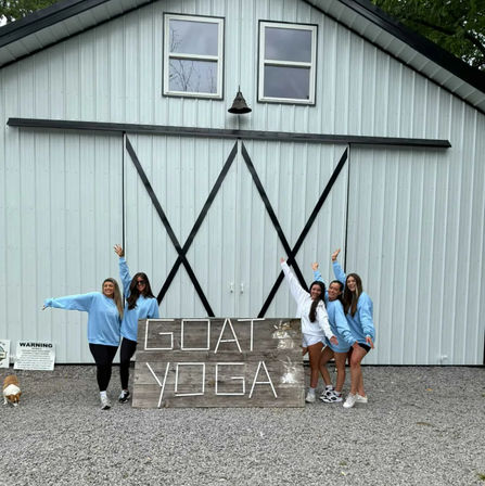 Five people in light-blue sweatshirts pose in front of a large white metal barn with black X trim and a rustic wooden sign reading GOAT YOGA, a small goat wanders on gravel at the left.