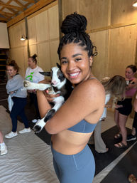 Smiling woman in blue activewear holding a black-and-white baby goat inside a wooden barn-style studio during a goat yoga session, people and mats visible in the background.