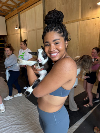 Smiling woman in blue activewear holding a black-and-white baby goat inside a wooden barn-style studio during a goat yoga session, people and mats visible in the background.