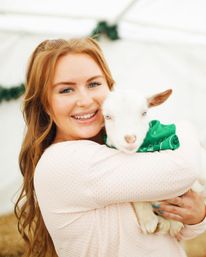 Smiling woman with long red hair hugging a white baby goat wearing a green bandana at a petting zoo inside a bright tent