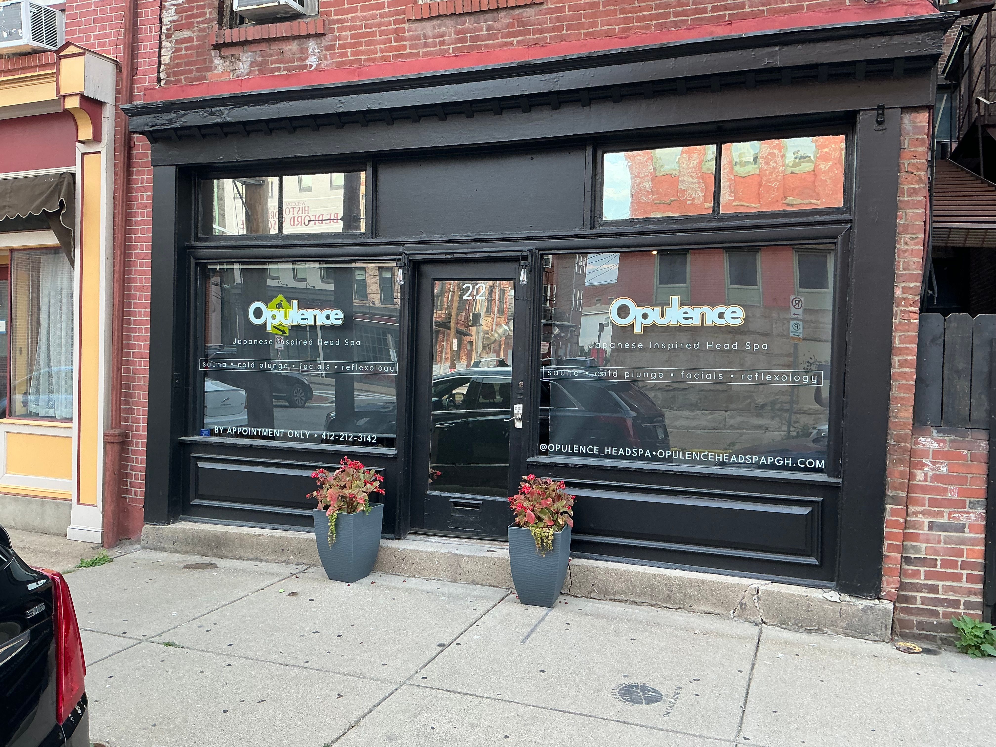 Black-painted storefront of a Japanese-inspired head spa in a red-brick building on an urban sidewalk, with two potted planters flanking the entrance and neighboring buildings reflected in the windows.