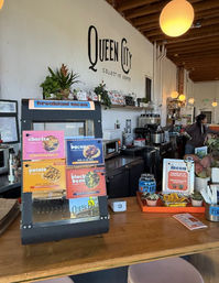 Cozy rustic coffee shop interior with wooden counter, colorful menu card display, espresso machines, pastries and potted plants beneath exposed beams and warm globe pendant lights.
