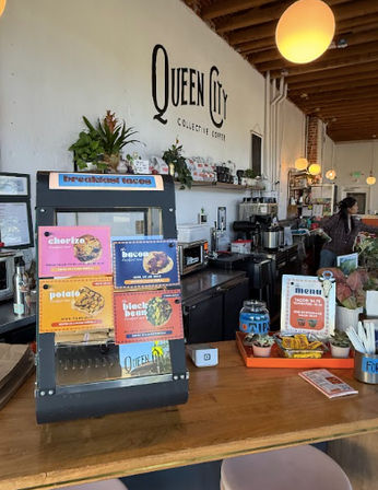 Cozy rustic coffee shop interior with wooden counter, colorful menu card display, espresso machines, pastries and potted plants beneath exposed beams and warm globe pendant lights.