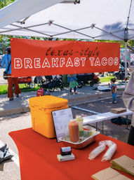 Outdoor food stall with a red "Texas-style BREAKFAST TACOS" banner under a white canopy; orange cooler, condiment bottles and wrapped tortillas on a red table at a sunny street market