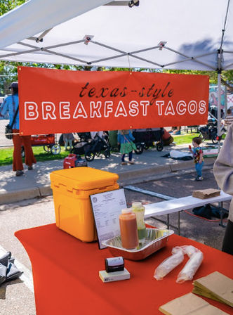 Outdoor food stall with a red "Texas-style BREAKFAST TACOS" banner under a white canopy; orange cooler, condiment bottles and wrapped tortillas on a red table at a sunny street market