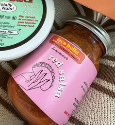 Glass mason jar of homemade red salsa with a bright pink label, tilted on a beige striped knit cloth beside a round dip container lid with green label — kitchen pantry-style product shot, ready for dipping.