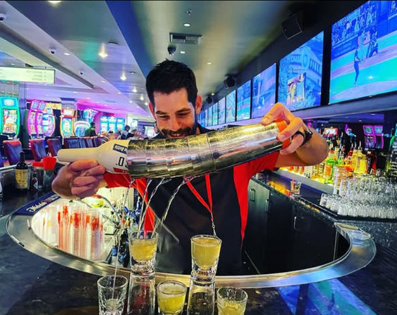 Smiling bartender performing a long-pour cocktail trick, pouring multiple yellow shots from a metal shaker across a curved casino bar with neon-lit slot machines and sports screens in the background.