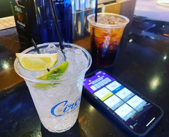 Two plastic bar drinks on a glossy counter — a sparkling lime cocktail with crushed ice and lime wedges in front and a dark iced mixed drink behind, next to a smartphone displaying a colorful app screen.