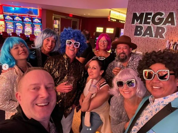 Group selfie of friends in colorful wigs, novelty sunglasses and sparkly outfits at a casino bar with slot machines and neon lighting, holding drinks and smiling.