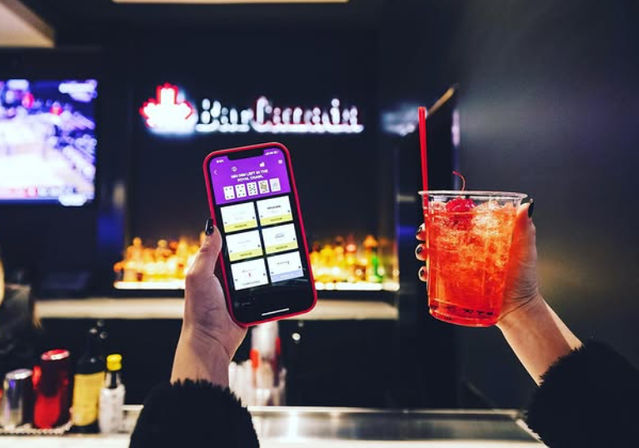 Hands holding a smartphone displaying a purple app and a red cocktail with straw at a dimly lit bar counter, with blurred neon sign and bottles in the background.
