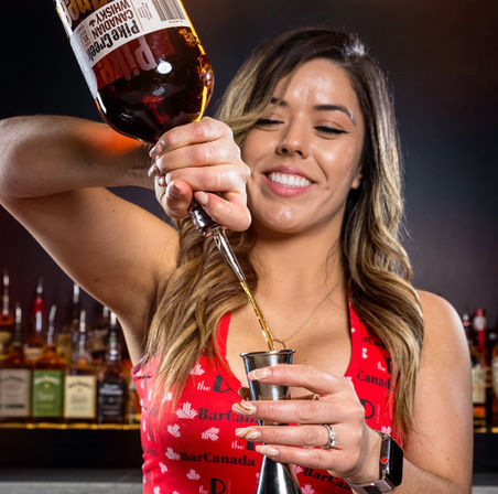 Cheerful bartender in a red top pouring amber whiskey from a bottle into a metal jigger at a bar with liquor shelves in the background