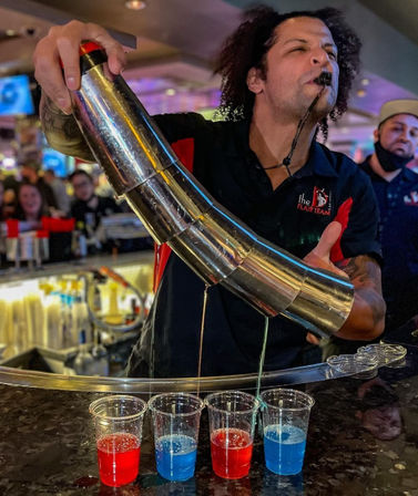 Flair bartender pouring blue and red layered shots from stacked metal shakers into clear plastic cups at a lively, crowded bar