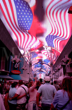 Crowd looking up at a giant LED canopy in downtown Las Vegas displaying a wavy American flag animation over a busy pedestrian street at night.