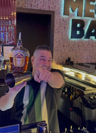 Smiling bartender behind a modern bar counter holding a liquor bottle and metal shaker while playfully pointing at the camera, with illuminated bottle shelves and a neon bar sign in the background.