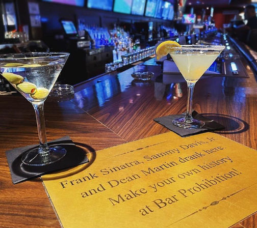 Two cocktails — a martini with olives and a lemon-garnished martini — on black napkins atop a glossy wooden bar next to a gold placard inviting patrons to make their own history, with blue-lit bottles and TV screens blurred in the background.